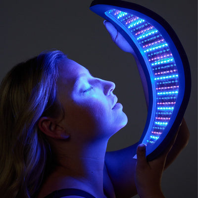 Person holding a crescent-shaped blue and red light therapy LED panel in front of their face against a dark background.
